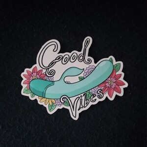 May include: A sticker with the words "Good Vibes" in a script font, featuring a teal vibrator design surrounded by pink, purple, and yellow flowers. The sticker has a white border and is on a black background.
