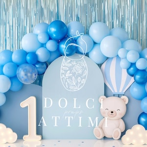 Può includere: Decorazione per festa di compleanno con arco di palloncini blu e bianchi, orsetto, numero 1 e luci a forma di nuvola. Lo sfondo include un pannello azzurro con la scritta "DOLCI ATTIMI".