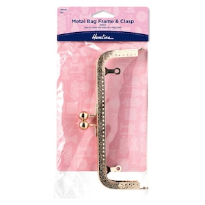 May include: A gold-colored metal bag frame and clasp, measuring 180mm, packaged in a clear plastic bag. The frame has a decorative pattern and includes links for a bag chain. The packaging features the text "Metal Bag Frame & Clasp" and the brand name "Hemline."