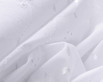 May include: A white fabric featuring delicate eyelet embroidery with small circular cutouts and swirling stitched patterns. The material is folded, creating soft shadows and highlights that emphasize the intricate needlework texture.
