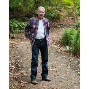 Puede incluir: Un hombre con una camisa de cuadros rojos y azules sobre una camiseta blanca, pantalones vaqueros azules y zapatos marrones. Está de pie en un camino en un bosque.