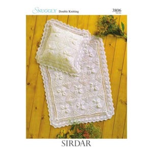 May include: A white knitted baby blanket and pillow set with a floral pattern. The set is made with a double knitting technique and features a delicate lace edging. The set is photographed on a wooden surface.