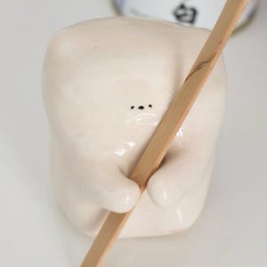 以下が含まれることがあります： 2つの点のような目と小さな口を持つシンプルな顔のオフホワイトの陶器製フィギュアが、長い薄茶色の竹製の茶杓を持っています。背景には、日本語の文字が書かれた円筒形の容器が一部見えています。