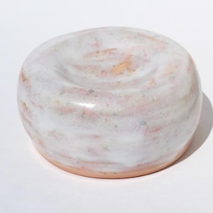 May include: A round ceramic dish with a smooth, glossy white glaze featuring subtle pink and green speckled accents. The object has a slightly concave centre and a thick, rounded rim, with the unglazed terracotta-coloured base visible at the bottom.