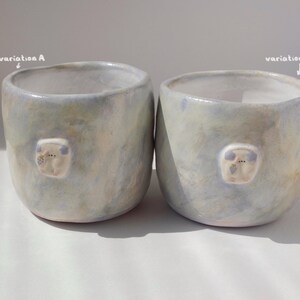 May include: Two handmade ceramic mugs with a light blue and cream glaze. Each mug features a small, raised, white and blue decorative element. The mugs are cylindrical with a slightly irregular shape, and the text "variation A" and "variation B" are visible.