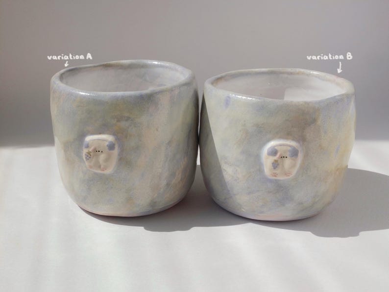 May include: Two handmade ceramic mugs with a light blue and cream glaze. Each mug features a small, raised, white and blue decorative element. The mugs are cylindrical with a slightly irregular shape, and the text "variation A" and "variation B" are visible.