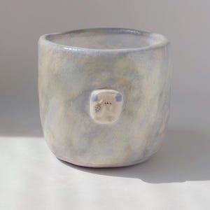 May include: A handmade ceramic mug with a light blue and cream mottled glaze. A small, square, white applique with a simple face is affixed to the front of the mug. The mug has a rounded shape and a smooth, unglazed rim.