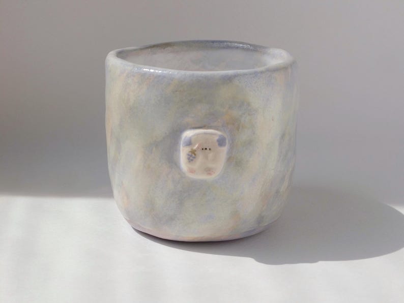 May include: A handmade ceramic mug with a light blue and cream mottled glaze. A small, square, white applique with a simple face is affixed to the front of the mug. The mug has a rounded shape and a smooth, unglazed rim.