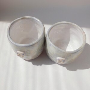 May include: Two handmade ceramic cups with a light blue and white glaze. Each cup has a small, square-shaped detail on the side. The cups are round with a wide opening and sit on a white surface.