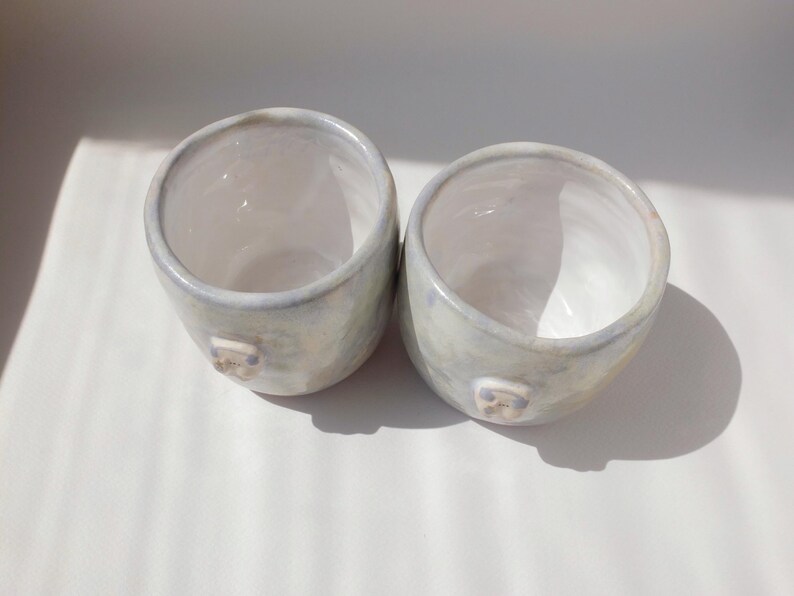 May include: Two handmade ceramic cups with a light blue and white glaze. Each cup has a small, square-shaped detail on the side. The cups are round with a wide opening and sit on a white surface.