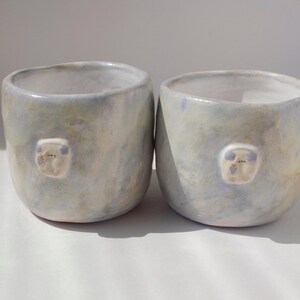 May include: Two handmade ceramic cups with a light blue and cream glaze. Each cup features a small, raised, white, bear-like face. The cups have a rounded shape and are suitable for beverages.