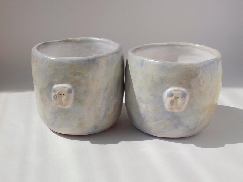May include: Two handmade ceramic cups with a light blue and cream glaze. Each cup features a small, raised, white, bear-like face. The cups have a rounded shape and are suitable for beverages.