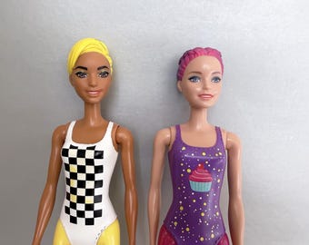 May include: Two dolls with distinct features. The left doll has short yellow hair and a white leotard with a black and white checkered pattern. The right doll has pink hair and a purple leotard featuring a cupcake graphic and yellow dots.
