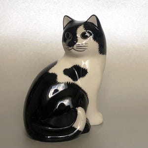 Könnte beinhalten: Eine Keramik-Katzenfigur in sitzender Position, mit glänzendem Schwarz-Weiß-Design. Die Katze hat schwarze Ohren, einen schwarzen Fleck um ein Auge und einen schwarzen Fleck an der Seite. Ein dekorativer Artikel.