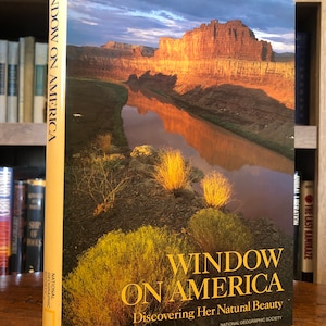 May include: A book titled "Window on America: Discovering Her Natural Beauty" by the National Geographic Society. The cover features a scenic landscape with a river flowing through a canyon, with red rock cliffs and a blue sky with clouds.