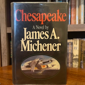 May include: A black hardback book with a red and white title, "Chesapeake", and the author's name, "James A. Michener". The cover features a painting of geese flying over a body of water.