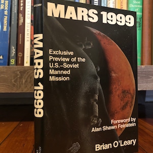 Op de afbeelding: Een zwart-wit boek getiteld "Mars 1999: Exclusieve preview van de bemande Amerikaans-Sovjetmissie" door Brian O'Leary met een voorwoord van Alan Shawn Feinstein. De omslag toont een zwart-wit illustratie van een ruimtevaartuig dat een rode planeet omcirkelt.
