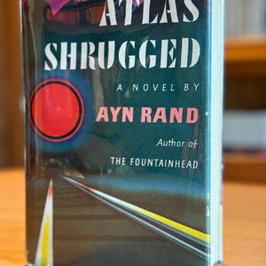 Pode incluir: Um livro de capa dura intitulado "Atlas Shrugged" de Ayn Rand. A capa apresenta um círculo vermelho, um design de estrada e o nome do autor em branco e vermelho. O livro está envolto em uma capa protetora transparente.