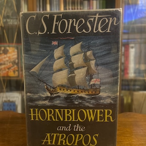 Puede incluir: Una cubierta de libro vintage con un barco con velas contra un mar azul oscuro. El título dice "Hornblower y el Atropos" por C.S. Forester.