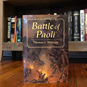 Op de afbeelding: Een gebonden boek getiteld "Battle of Paoli" door Thomas J. McGuire. De omslag toont een schilderij van een veldslag met rook en vuur.