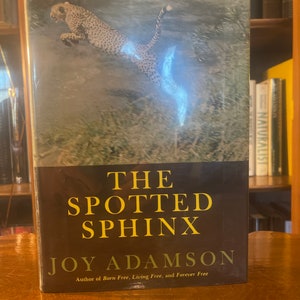 May include: A book cover with a cheetah jumping in the air. The title of the book is "The Spotted Sphinx" by Joy Adamson. The book cover is black with gold lettering.