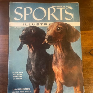 May include: A vintage Sports Illustrated magazine cover featuring two Dachshunds, one black and one brown, against a light blue background. The cover text reads "Sports Illustrated" and "December 12, 1955".