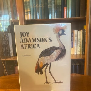 May include: A book titled "Joy Adamson's Africa" with a picture of a grey crowned crane on the cover. The crane is standing on one leg with its head held high. The book is on a wooden table.
