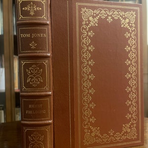 Puede incluir: Un libro encuadernado en cuero marrón con letras doradas y diseños decorativos. El título del libro es "TOM JONES" de "HENRY FIELDING".
