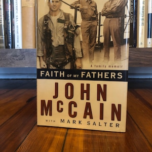 May include: A book titled "Faith of My Fathers" by John McCain with Mark Salter. The cover features a black and white photograph of three men in military uniforms. The book is a family memoir.