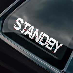 May include: A black car with a white "STANDBY" sticker on the rear window. The sticker has a glitch effect with red and blue lines. A round sticker with the text "BAD INVESTMENTS AUTO 2016" is on the side of the car.