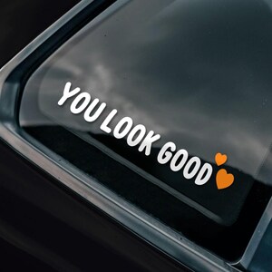 May include: A black car with a white vinyl sticker on the rear window that says "You Look Good" with two orange hearts. The sticker is partially peeled back to reveal a black and white sticker underneath that says "Bad Investments Auto 2016" with a stylized letter "A" in the centre.