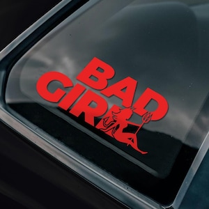 May include: Red vinyl decal with the words "Bad Girl" and a silhouette of a woman with a trident. The decal is on the back window of a black car.