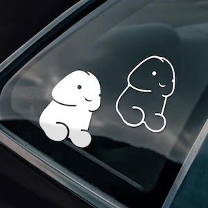 May include: Two white vinyl car stickers showing cartoon penises with smiling faces. One is a solid white shape, the other an outline. The stickers are applied to a car window.