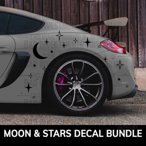 May include: A grey car with a black moon and stars decal on the back. The decal is on the rear quarter panel and includes a crescent moon and several stars.