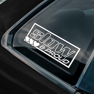 May include: White rectangular sticker with the words "SLOW & PROUD" in a bold, outlined font. Three white hearts are on the left side. The sticker is affixed to a car window, with a dark background.