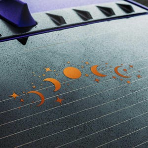May include: Orange moon phase and star decals on a car window. The design features a series of crescent moons, a full moon, and stars in various sizes. The decals are arranged in a horizontal line.