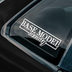 May include: White vinyl decal with the words "BASE MODEL" in a rectangular frame, and the word "beauty" in cursive script. The decal is affixed to the rear window of a black vehicle.