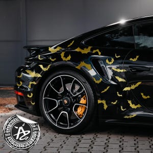 May include: Black sports car adorned with gold bat decals. The car features black alloy wheels with white detailing and orange brake calipers. A sticker with the text "BAD INVESTMENTS AUTO" is visible.