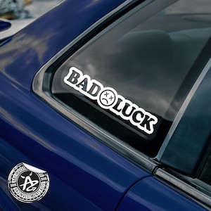 May include: A white vinyl sticker with the text "BAD LUCK" in a bold, rounded font. A black and white cartoon face with an X for eyes is included in the text. The sticker is on the back window of a blue car.