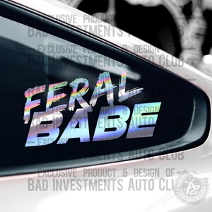 May include: A holographic sticker with the text "FERAL BABE" in a bold, white font with a rainbow iridescent background. The sticker is on a black car window. The text "EXCLUSIVE PRODUCT & DESIGN OF BAD INVESTMENTS AUTO CLUB" is visible at the bottom of the image.