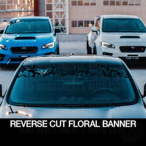 May include: A black car with a floral windshield banner that reads "Bad Investments Auto Club". The car is parked in front of two other cars, one blue and one white.