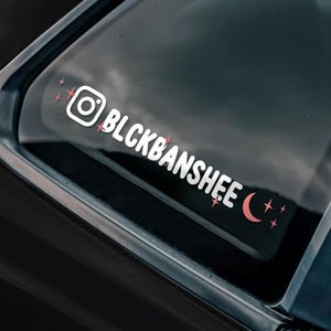May include: White vinyl sticker with the text "BLCKBANSHIEE" and an Instagram icon, a crescent moon, and stars in pink.