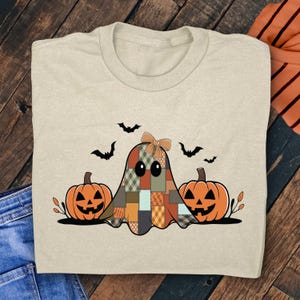 May include: Beige t-shirt featuring a patchwork ghost design with a bow, two jack-o'-lanterns, and three bats. The ghost has a variety of patterned squares in shades of orange, brown, and green. The pumpkins are orange with carved faces.
