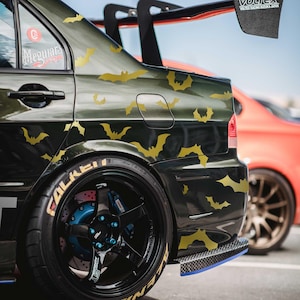 May include: Dark green car with gold bat decals. The car has black rims with gold lettering and blue brake calipers. A red car is partially visible in the background. The car has a black spoiler.