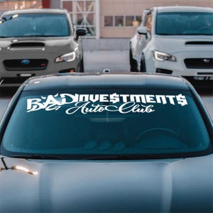 May include: A close-up of a car's windshield with a white decal that reads "BAD INVESTMENTS Auto Club" in a stylized font. The car is gray, and two other cars are visible in the background.
