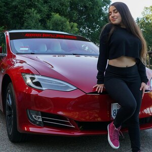May include: A red Tesla Model S with a black windshield banner that reads "Game for the long run". A person is sitting on the hood of the car.