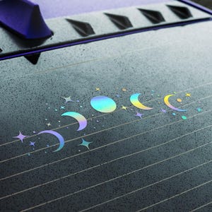 May include: A car window with a holographic moon phases sticker. The sticker features a full moon, crescent moons, and stars in a rainbow iridescent colour.