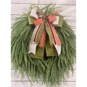 May include: A green fern wreath with a large bow. The bow features olive green, striped, and coral-colored ribbons. The wreath is made of faux greenery and is suitable for indoor or outdoor use.
