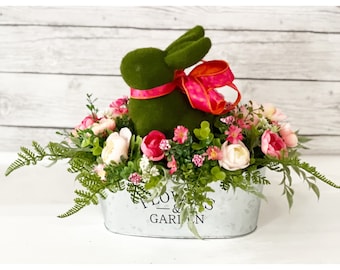 May include: A decorative Easter centerpiece featuring a green bunny with a pink ribbon, surrounded by pink and white flowers and greenery. The arrangement is in a metal container with the words "FLOWERS & GARDEN".