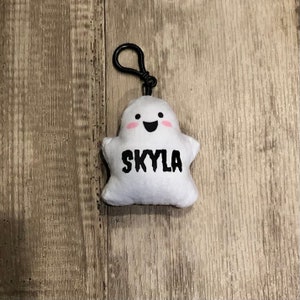 May include: White plush ghost keychain with a black clip. The ghost has a smiling face with pink cheeks and the name "Skyla" written in black.